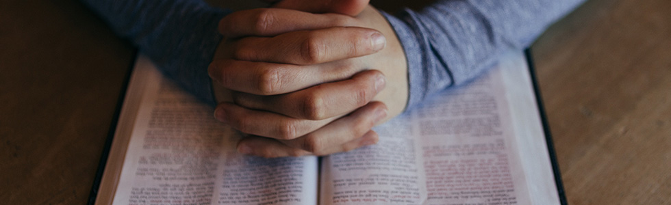 A person's folded hands praying on top of the Bible
